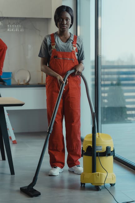 A professional cleaner, dressed in grey top and red overalls, operates a yellow industrial vacuum cleaner in a modern kitchen with a light grey tiled floor, white cabinets, and a minimalist design. The cleaner is standing upright near a glass sliding door, which lets in natural daylight, highlighting the clean and shiny surface of the floor. The kitchen appears freshly cleaned, with no visible dust or debris, emphasizing surface cleaning and maintenance. The image showcases domestic cleaning activities, aligning with the services offered by Carpet Cleaners Kingston, and demonstrates deep cleaning and sanitisation procedures in a residential setting.