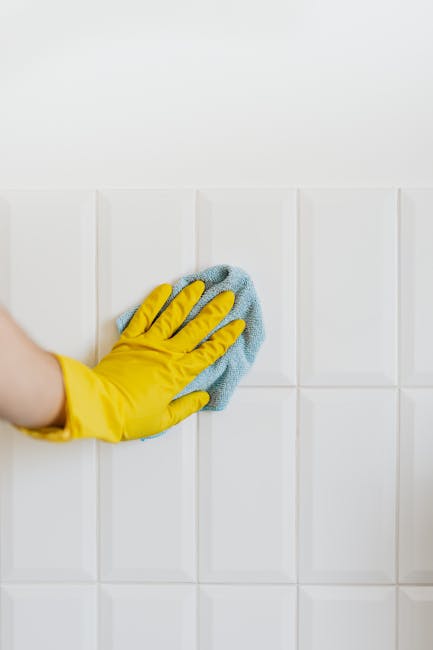 Close-up of a person wearing a yellow rubber cleaning glove using a light blue microfiber cloth to clean white ceramic tiled wall in a kitchen or bathroom. The tiles are smooth and glossy, reflecting light, and appear freshly cleaned with no visible dust or stains. The cloth is folded over the hand, and the cleaning process is focused on the surface. The background shows more tiles with a consistent pattern and neat grout lines, under bright, natural lighting. This image demonstrates surface cleaning and sanitisation, emphasizing thorough domestic or commercial cleaning services provided by Carpet Cleaners Kingston, as referenced in the page titled 'Avoid hidden cleaning charges in Kingston home cleans, KINGSTON'.