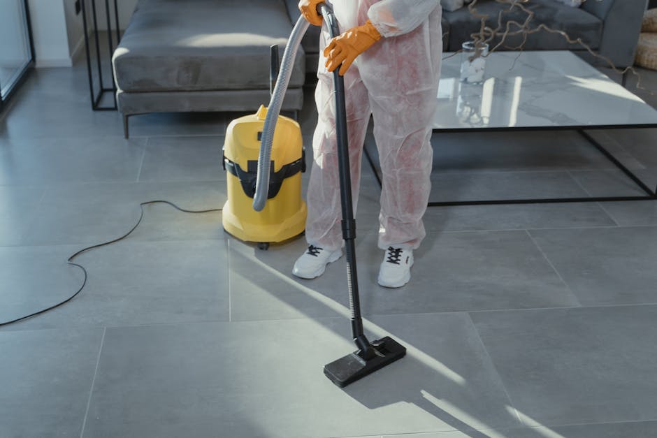 A professional cleaner, dressed in grey top and red overalls, operates a yellow industrial vacuum cleaner in a modern kitchen with a light grey tiled floor, white cabinets, and a minimalist design. The cleaner is standing upright near a glass sliding door, which lets in natural daylight, highlighting the clean and shiny surface of the floor. The kitchen appears freshly cleaned, with no visible dust or debris, emphasizing surface cleaning and maintenance. The image showcases domestic cleaning activities, aligning with the services offered by Carpet Cleaners Kingston, and demonstrates deep cleaning and sanitisation procedures in a residential setting.