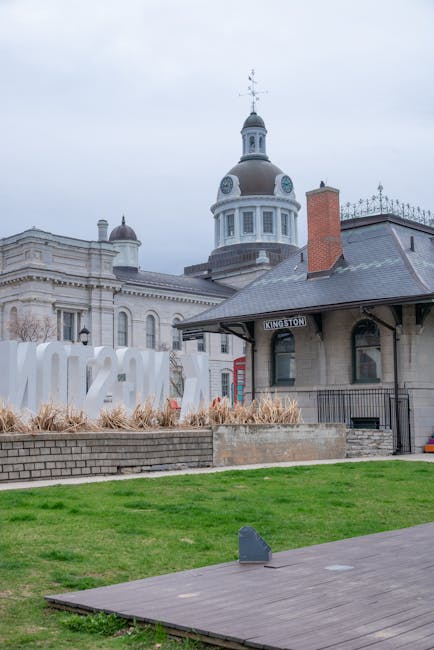 Exterior view of the Bentall Centre in Kingston, showing a historic building with a domed clock tower and classical architectural details. In the foreground, there is a manicured green lawn with a paved walkway and a small rectangular grey object, possibly a security or surveillance device. The sky is overcast, casting diffuse light over the scene. Visible nearby is a modern wooden deck area, indicating a space for leisure or waiting. The overall setting emphasizes a clean and maintained urban environment, relevant to surface cleaning and maintenance in commercial premises, as highlighted in the Kingston area, where Carpet Cleaners Kingston provides professional cleaning services for shop owners.