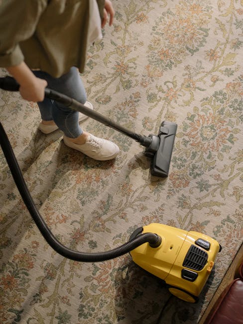 A person using a black vacuum cleaner to clean a traditional patterned area rug with floral motifs in a well-lit indoor space. The vacuum head is flat on the surface, with visible dust and debris being removed, and the user is wearing casual clothing and white sneakers. The room features a wooden floor and natural lighting that highlights the clean, well-maintained appearance of the rug, as part of a comprehensive surface cleaning and sanitisation process conducted by Carpet Cleaners Kingston, tailored for shop owners at the Bentall Centre Kingston, KINGSTON.