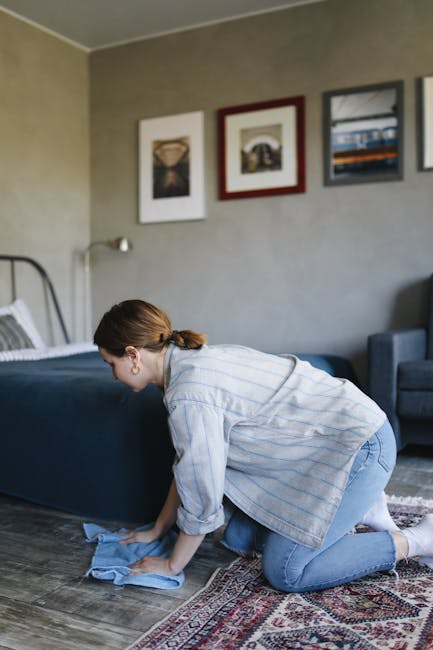 A woman with curly hair, dressed in a beige jacket, blue jeans, and white shoes, is performing surface cleaning with a vacuum cleaner on a patterned rug in a living room. She is holding the vacuum's hose and nozzle, focusing on the area around a maroon leather sofa. The room features wooden flooring and a wooden sideboard with decorative items, including a small statue and framed pictures, in the background. The space is well-lit with natural light, emphasizing the cleanliness and freshness of the carpet and surrounding surfaces. This deep cleaning process, carried out by Carpet Cleaners Kingston, highlights their expertise in domestic cleaning and rug maintenance for riverside homes in Kingston.