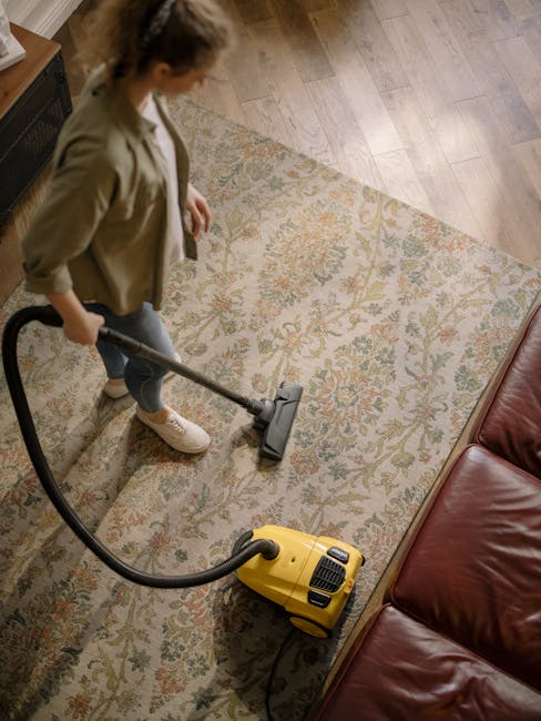 A person using a vacuum cleaner on a patterned area rug in a living room setting, with visible parts of their legs, white shoes, and beige pants. The vacuum cleaner has a yellow body and a black flexi