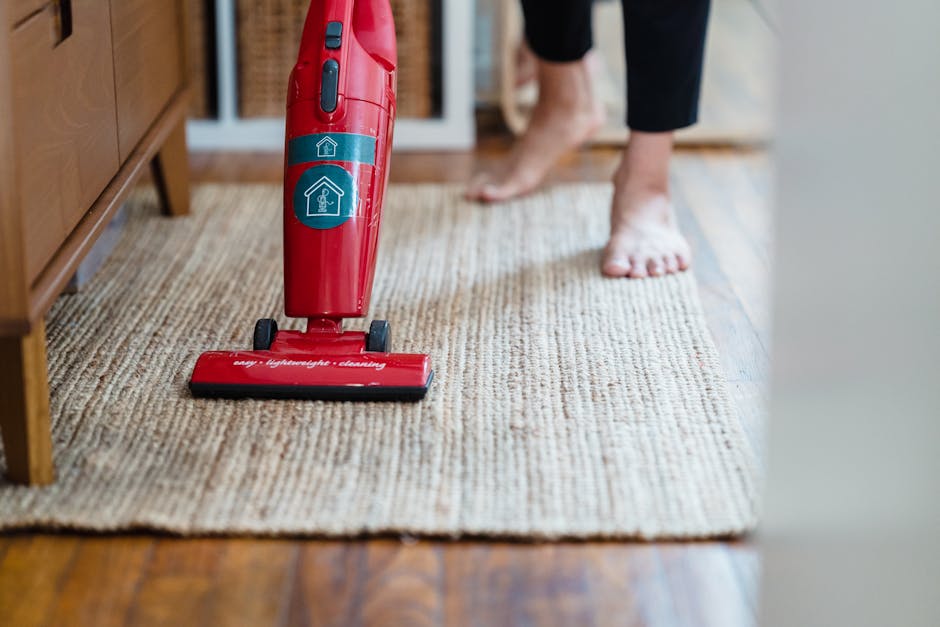 Close-up of a red handheld vacuum cleaner with dual buttons on the handle, positioned on a beige woven area rug in a home setting. The vacuum is actively being used for surface cleaning, focusing on the rug with a wooden floor visible at the edge. In the background, a person wearing black pants and barefoot is standing on the rug, with their feet partially visible. The room features natural light, likely from a nearby window, illuminating the scene. The setting appears to be a living room or corridor area, emphasizing the importance of regular deep cleaning and maintenance to avoid common carpet cleaning mistakes. The image highlights the tools and cleanliness condition associated with domestic cleaning, demonstrating effective surface sanitisation practices that Carpet Cleaners Kingston advocates.