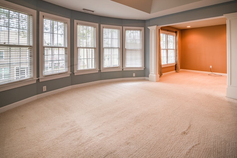 A spacious, well-lit living room with a beige carpet that appears clean and plush, stretching across the entire floor area. The room features a curved wall with six large windows fitted with white blinds, allowing natural light to brighten the space. Adjacent to this bay window area is another section of the room with a large window adorned with light brown curtains, providing additional light and a cozy atmosphere. The walls are painted in a soft gray tone, complementing the white crown molding and baseboards that frame the room. The carpet, in good condition, is free of stains or debris, highlighting proper surface cleaning and maintenance. Visible elements include a single electrical outlet on the wall, and the room is illuminated by natural daylight, creating a clean and inviting environment. This setting demonstrates the importance of regular deep cleaning and surface maintenance, aligning with best practices in domestic cleaning as promoted by Carpet Cleaners Kingston, especially for landlords seeking to avoid common carpet cleaning mistakes.