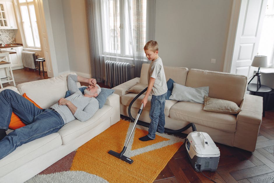 Close-up of a red handheld vacuum cleaner with dual buttons on the handle, positioned on a beige woven area rug in a home setting. The vacuum is actively being used for surface cleaning, focusing on the rug with a wooden floor visible at the edge. In the background, a person wearing black pants and barefoot is standing on the rug, with their feet partially visible. The room features natural light, likely from a nearby window, illuminating the scene. The setting appears to be a living room or corridor area, emphasizing the importance of regular deep cleaning and maintenance to avoid common carpet cleaning mistakes. The image highlights the tools and cleanliness condition associated with domestic cleaning, demonstrating effective surface sanitisation practices that Carpet Cleaners Kingston advocates.