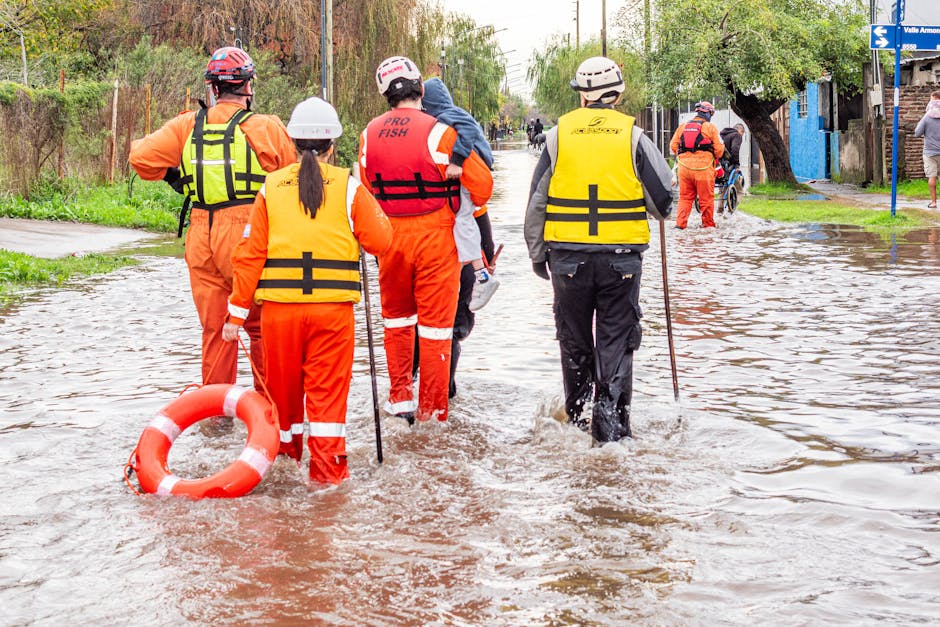 A team of emergency responders wearing orange and yellow waterproof suits, helmets, and life vests wading through a flooded street with muddy water. They are equipped with safety gear including poles and a life ring, assessing the flood situation amid submerged sidewalks and partially visible trees and buildings in the background. The scene is outdoors in a residential area during daylight, with overcast weather conditions. This image illustrates flood response efforts related to emergency management and surface water safety, aligning with the urgent flood response services offered by Carpet Cleaners Kingston for surface cleaning and flood damage recovery.