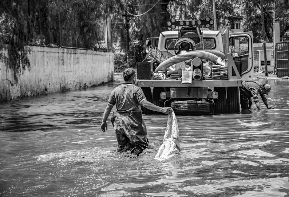In a black-and-white photograph, two workers are engaged in emergency flood response, demonstrating surface cleaning and removal of floodwaters from a residential street. The scene shows one man in the foreground wearing a rubber apron and boots, holding a cloth or tarp while standing in waist-high water, with a determined expression. In the background, another worker is bent over near a large flood pump truck, which is equipped with hoses and machinery for water extraction and deep cleaning. The environment appears to be an outdoor area with a concrete wall and fencing visible, and the lighting highlights the reflective, wet surfaces of the floodwater, emphasizing their cleaning efforts during this emergency response. This image illustrates the active process of flood mitigation and surface sanitisation, which aligns with the urgent services offered by Carpet Cleaners Kingston in their cleaning and disaster recovery campaigns.