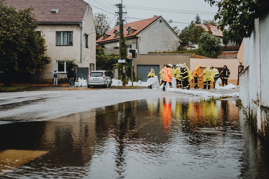 A team of emergency responders wearing orange and yellow waterproof suits, helmets, and life vests wading through a flooded street with muddy water. They are equipped with safety gear including poles and a life ring, assessing the flood situation amid submerged sidewalks and partially visible trees and buildings in the background. The scene is outdoors in a residential area during daylight, with overcast weather conditions. This image illustrates flood response efforts related to emergency management and surface water safety, aligning with the urgent flood response services offered by Carpet Cleaners Kingston for surface cleaning and flood damage recovery.