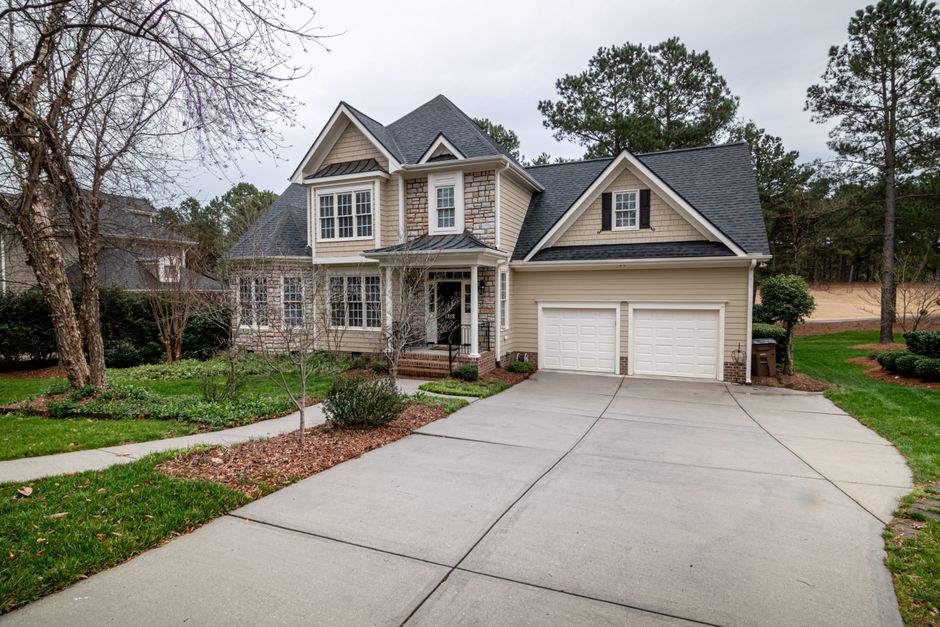 A modern two-story house with a well-maintained concrete driveway leading to a double garage, surrounded by landscaped gardens with grass, small trees, and shrubs. The house features beige siding with white trim, multiple gabled roofs covered with dark shingles, and large windows with white frames. An entrance with brick steps and a small porch is visible, with a pathway curving through the front yard. The driveway appears clean and dry, with no visible stains or moisture, and the overall scene suggests a tidy, residential setting. The image highlights aspects of surface cleaning and maintenance in a domestic environment, consistent with the services offered by Carpet Cleaners Kingston, especially in relation to monitoring and preventing driveway damp issues caused by hidden problems when driveway cleaning is performed, as discussed in the page titled 'Hidden problems when driveway cleaning causes damp in Kingston'.