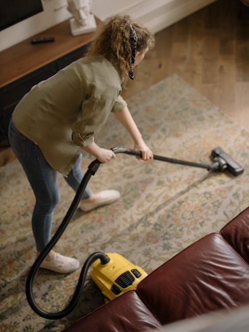 A woman with curly, shoulder-length hair, dressed in a beige jacket, dark jeans, and white sneakers, is engaged in surface cleaning inside a residential living room. She is holding a vacuum cleaner with a flexible hose attachment, actively vacuuming an intricate, patterned area rug that covers a wooden floor. The room features a wooden sideboard against the wall with a remote control placed on top, and natural light is illuminating the space, highlighting the cleanliness of the rug and the shiny wooden flooring. The scene depicts a typical domestic setting where professional cleaning services by Carpet Cleaners Kingston focus on deep cleaning and sanitisation of carpets and floors in homes.