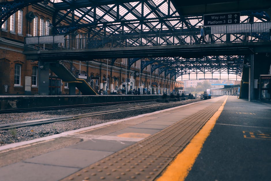A photograph of an outdoor railway station platform captures a wooden and concrete surface with a textured tactile paving strip along the edge for safety. The platform features blue-painted metal supp