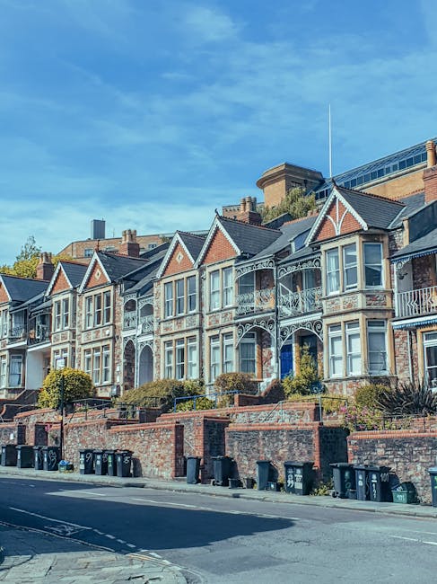 Photograph of a residential street with a row of multi-story brick buildings featuring large rectangular windows with white frames. The buildings have a classic Victorian style with decorative arches 
